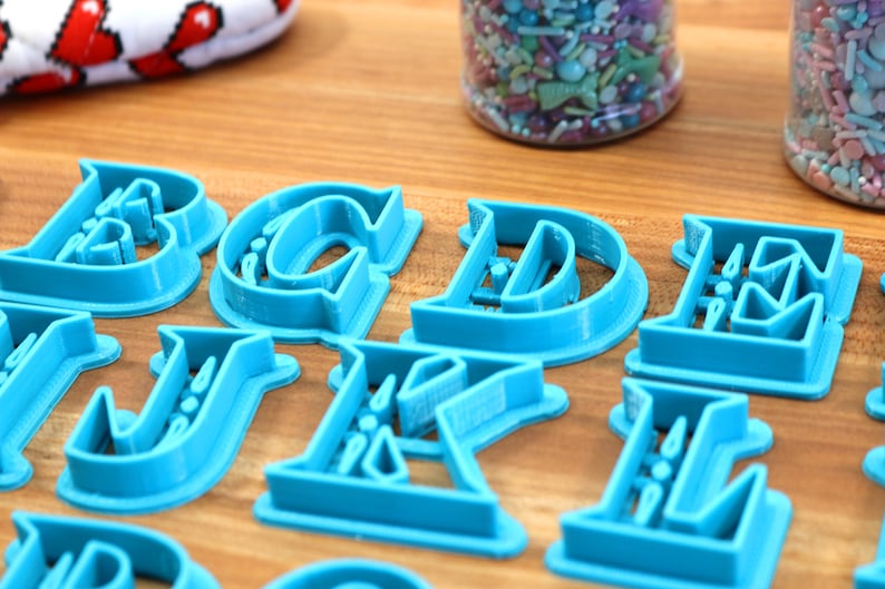 May include: A set of blue plastic alphabet cookie cutters. The cutters are arranged on a wooden surface.
