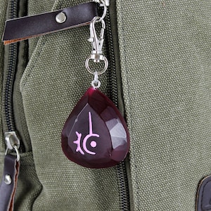 Op de afbeelding: Een donkergroene canvas rugzak met een zilveren rits en een rode en roze kristallen hanger bevestigd aan de rits. De hanger heeft een roze symbool erop.