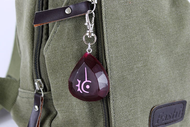 Op de afbeelding: Een donkergroene canvas rugzak met een zilveren rits en een rode en roze kristallen hanger bevestigd aan de rits. De hanger heeft een roze symbool erop.