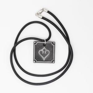 May include: A black cord necklace with a silver pendant featuring a stylized heart design. The pendant is square shaped with a raised border.