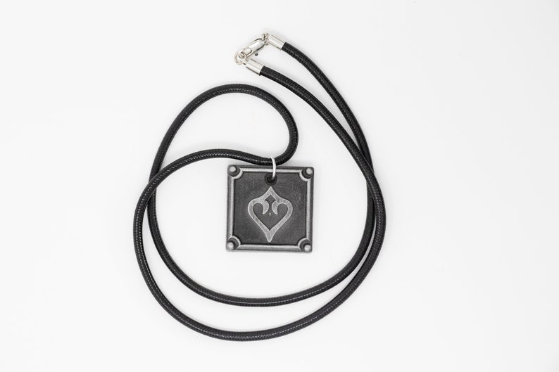 May include: A black cord necklace with a silver pendant featuring a stylized heart design. The pendant is square shaped with a raised border.
