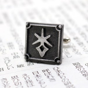 May include: Silver cufflinks with a square design featuring a stylized Japanese character in the center. The character is in a silver color against a black background.