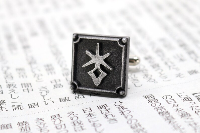 May include: Silver cufflinks with a square design featuring a stylized Japanese character in the center. The character is in a silver color against a black background.