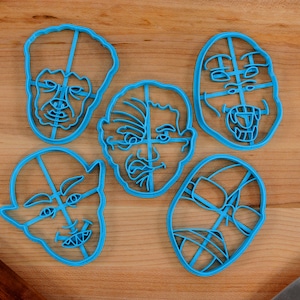 May include: Five blue plastic cookie cutters shaped like different faces. The faces are all different expressions, including a surprised face, a frowning face, and a face with its tongue sticking out.