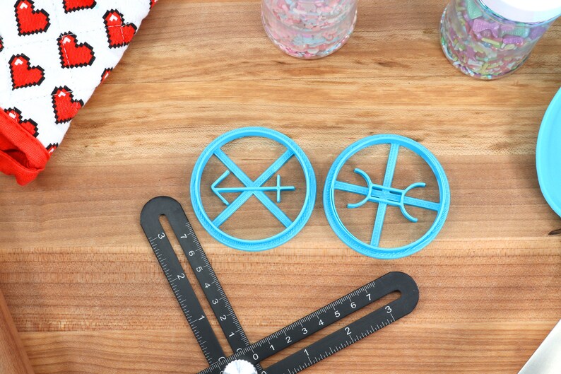 May include: Two blue plastic cookie cutters on a wooden surface. One cutter is shaped like a cross with an arrow pointing to the left. The other cutter is shaped like a circle with four spokes.
