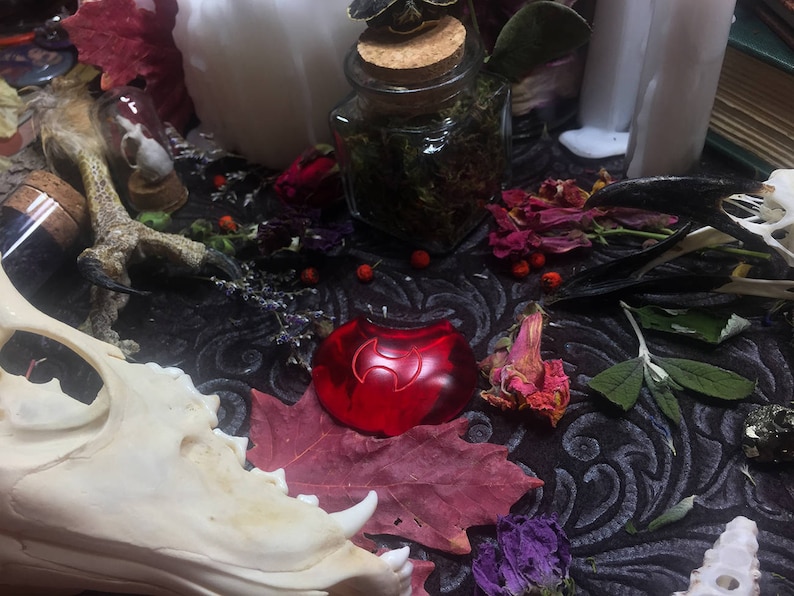 May include: A red glass crescent moon charm with a stylized symbol inside, surrounded by dried flowers, herbs, and animal bones on a black fabric background.