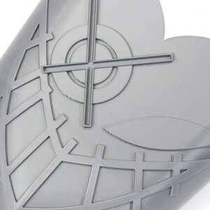 May include: A gray, heart-shaped object with a geometric design. The design includes a crosshair, a circle, and a grid-like pattern. The object has a smooth, slightly curved surface.
