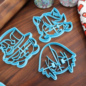 May include: Three blue plastic cookie cutters shaped like cartoon characters. The cutters are on a wooden surface.