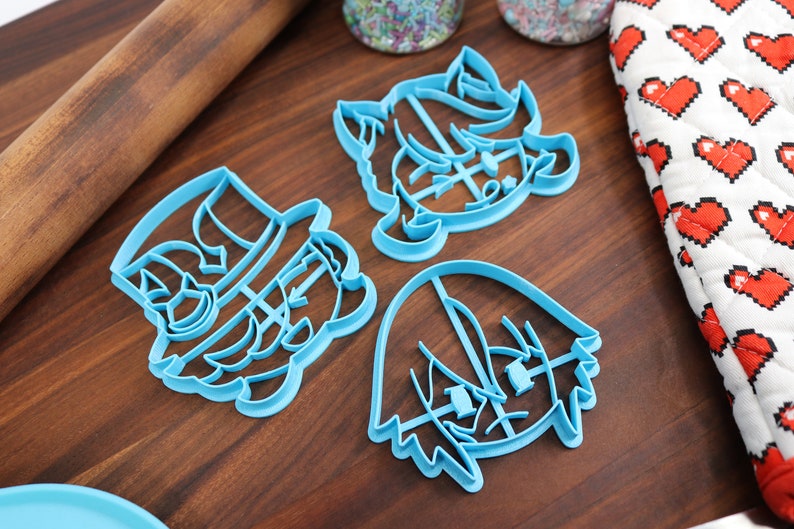 May include: Three blue plastic cookie cutters shaped like cartoon characters. The cutters are on a wooden surface.