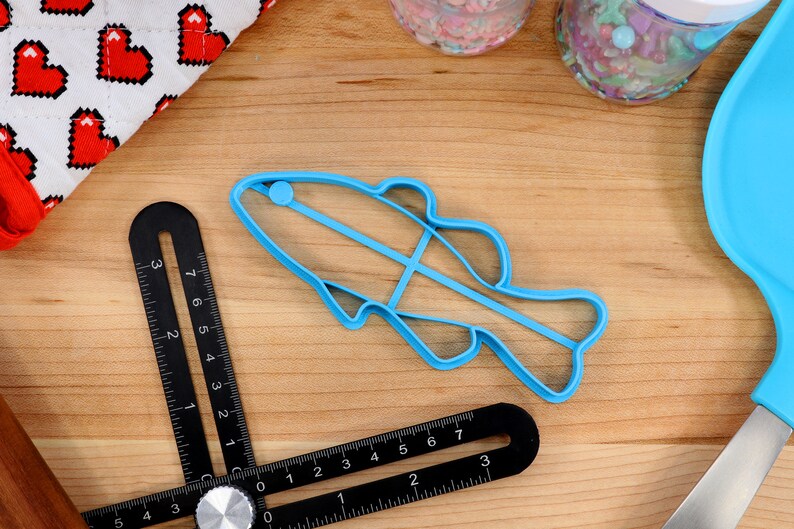 May include: A blue plastic fish-shaped cookie cutter on a wooden surface. The cutter is about 5 inches long and has a small blue circle on the top.