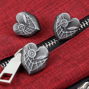 K&ouml;nnte beinhalten: Drei silberne herzf&ouml;rmige Pins mit einem Netzdesign und einem Ziel in der Mitte. Die Pins befinden sich auf einem roten Stoffhintergrund.