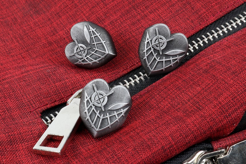 K&ouml;nnte beinhalten: Drei silberne herzf&ouml;rmige Pins mit einem Netzdesign und einem Ziel in der Mitte. Die Pins befinden sich auf einem roten Stoffhintergrund.
