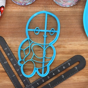 May include: A blue plastic cookie cutter shaped like a gingerbread person with a smiling face and a detailed design. The cutter is on a wooden surface.