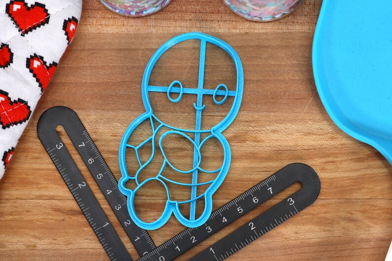 May include: A blue plastic cookie cutter shaped like a gingerbread person with a smiling face and a detailed design. The cutter is on a wooden surface.