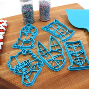 May include: A set of six blue plastic cookie cutters shaped like cartoon characters on a wooden surface. The cutters are arranged in a semi-circle with a spatula and a rolling pin in the background.