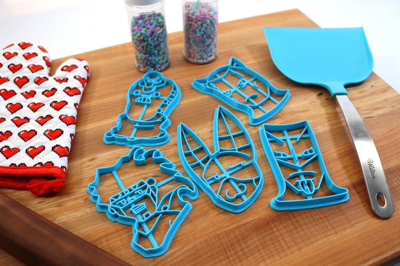 May include: A set of six blue plastic cookie cutters shaped like cartoon characters on a wooden surface. The cutters are arranged in a semi-circle with a spatula and a rolling pin in the background.