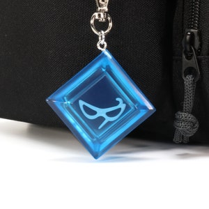 May include: A blue, diamond-shaped keychain with a white eye mask design. The keychain is attached to a silver metal chain.
