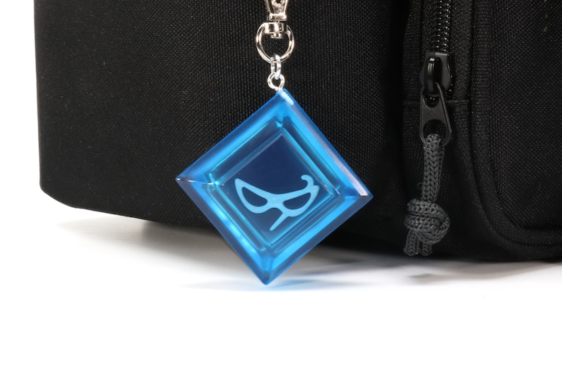 May include: A blue, diamond-shaped keychain with a white eye mask design. The keychain is attached to a silver metal chain.