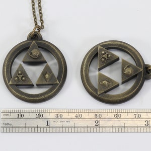 Puede incluir: Dos collares de color dorado con un colgante de Trifuerza. El colgante es de dise&ntilde;o circular con una estrella de tres puntas en el centro. La estrella est&aacute; formada por tres tri&aacute;ngulos, cada uno con un s&iacute;mbolo diferente. Los collares est&aacute;n sobre un fondo blanco con una regla para la escala.