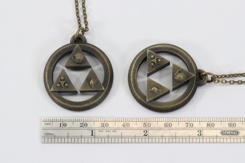 Puede incluir: Dos collares de color dorado con un colgante de Trifuerza. El colgante es de dise&ntilde;o circular con una estrella de tres puntas en el centro. La estrella est&aacute; formada por tres tri&aacute;ngulos, cada uno con un s&iacute;mbolo diferente. Los collares est&aacute;n sobre un fondo blanco con una regla para la escala.