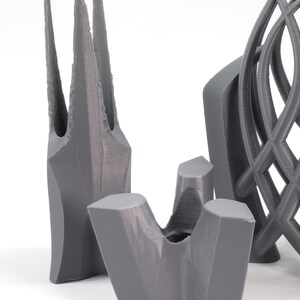 May include: Three gray 3D printed objects. The first object is tall and slender with a pointed top. The second object is a short, wide, and open-topped vase. The third object is a tall, intricate, and open-work sculpture.