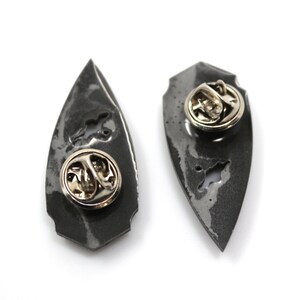 May include: Two silver-colored metal pins in the shape of a teardrop. The pins have a textured surface and a silver-colored pin back.