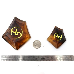 May include: Two brown resin dice with a gold symbol on each face. The dice are different sizes and are sitting on a white surface.