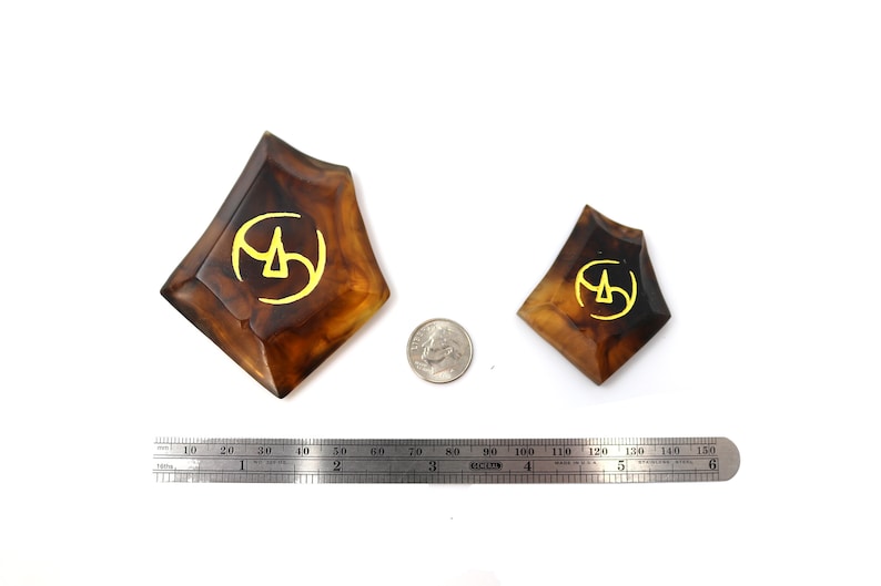 May include: Two brown resin dice with a gold symbol on each face. The dice are different sizes and are sitting on a white surface.