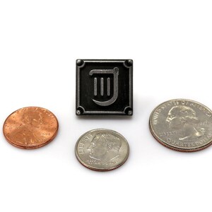 Puede incluir: Un pin de metal negro con un dise&ntilde;o de arpa plateado dentro de un marco cuadrado. El pin est&aacute; rodeado de tres monedas estadounidenses: un centavo, una moneda de diez centavos y una moneda de veinticinco centavos.