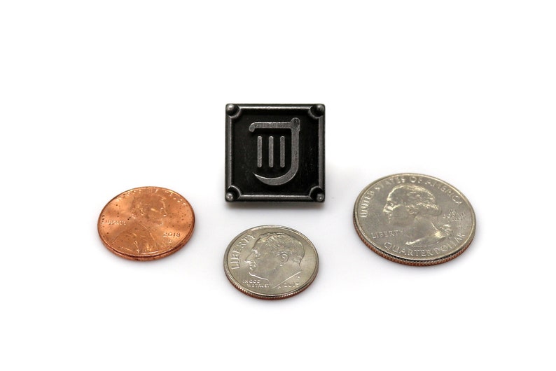 Puede incluir: Un pin de metal negro con un dise&ntilde;o de arpa plateado dentro de un marco cuadrado. El pin est&aacute; rodeado de tres monedas estadounidenses: un centavo, una moneda de diez centavos y una moneda de veinticinco centavos.