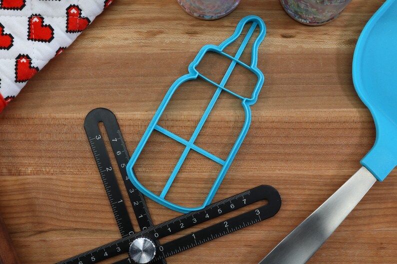 May include: A blue plastic baby bottle cookie cutter on a wooden surface. The cutter has a detailed design with a nipple and a handle.
