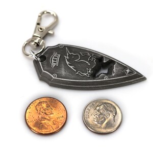 May include: A silver metal keychain shaped like a shield with a unicorn design. The keychain has a silver metal clasp and is next to two US coins, a penny and a dime.