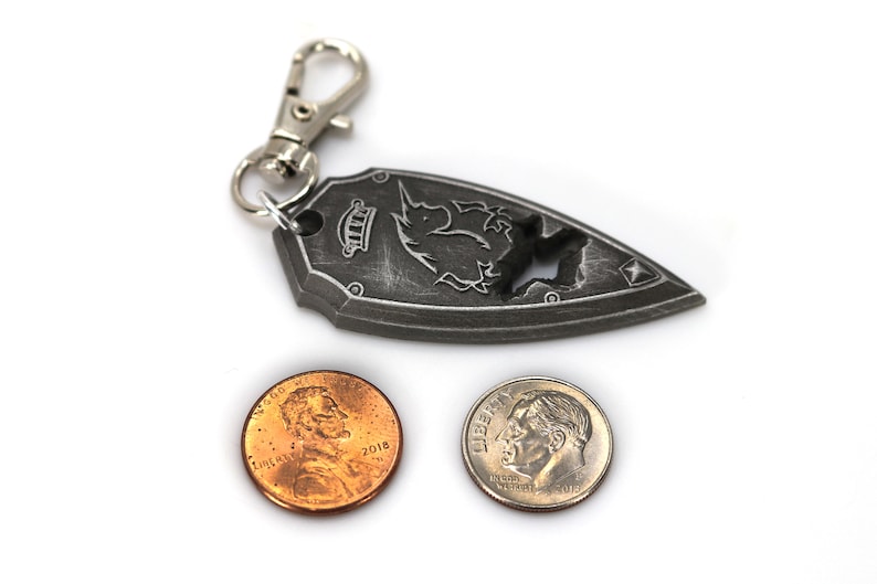 May include: A silver metal keychain shaped like a shield with a unicorn design. The keychain has a silver metal clasp and is next to two US coins, a penny and a dime.