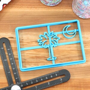 May include: Blue plastic cookie cutter shaped like the South Carolina state flag. The flag features a palmetto tree and crescent moon.