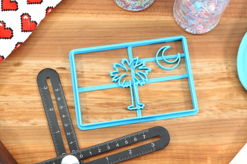 May include: Blue plastic cookie cutter shaped like the South Carolina state flag. The flag features a palmetto tree and crescent moon.