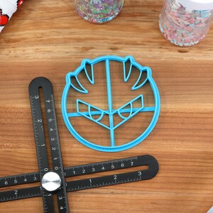 May include: Blue plastic cookie cutter shaped like a cartoon face with angry eyes and a wide mouth.