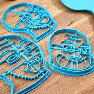 May include: Three blue plastic cookie cutters shaped like faces with detailed features, including eyes, noses, and mouths. The cutters are on a wooden surface.