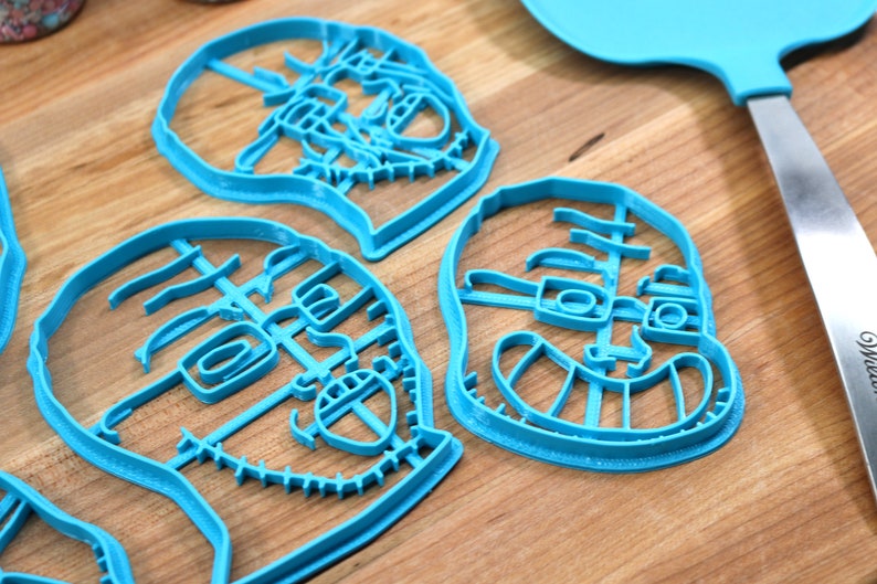 May include: Three blue plastic cookie cutters shaped like faces with detailed features, including eyes, noses, and mouths. The cutters are on a wooden surface.