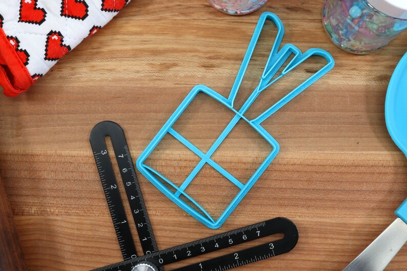 K&ouml;nnte beinhalten: Ein blauer Plastik-Ausstecher in Form einer Geschenkbox mit Schleife oben. Der Ausstecher liegt auf einer Holzoberfl&auml;che.