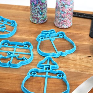 May include: A set of blue plastic cookie cutters shaped like frogs, flowers, and mushrooms. The cutters are on a wooden surface with jars of sprinkles and a blue spatula.