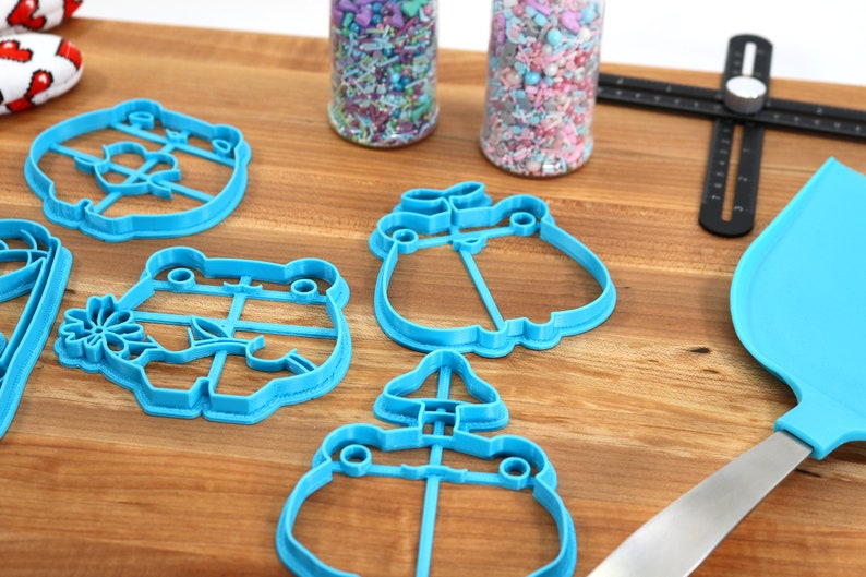 May include: A set of blue plastic cookie cutters shaped like frogs, flowers, and mushrooms. The cutters are on a wooden surface with jars of sprinkles and a blue spatula.