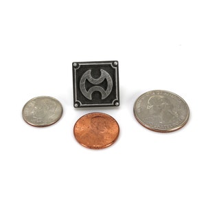 May include: A silver-coloured pin with a black square background and a stylised silver symbol. The pin is surrounded by three UK coins: a twenty pence piece, a one pence piece, and a ten pence piece.