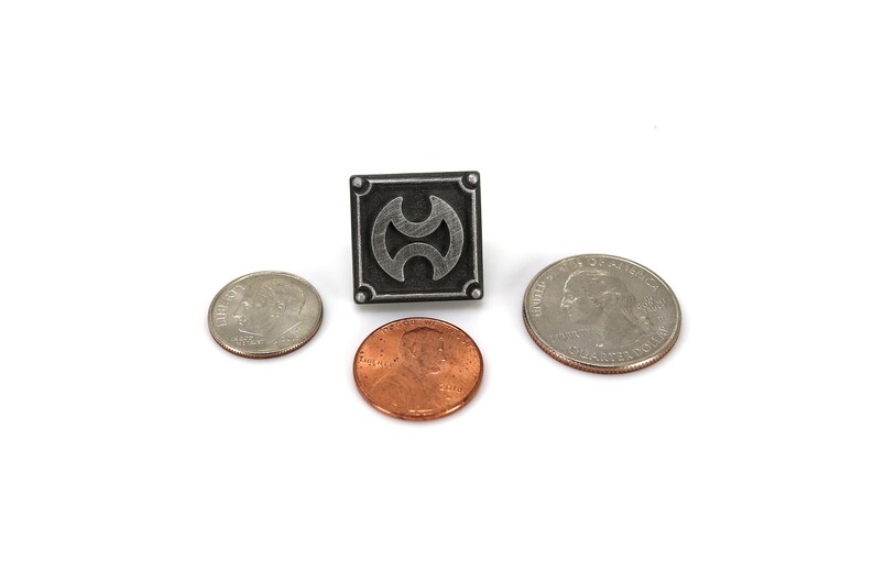 May include: A silver-coloured pin with a black square background and a stylised silver symbol. The pin is surrounded by three UK coins: a twenty pence piece, a one pence piece, and a ten pence piece.