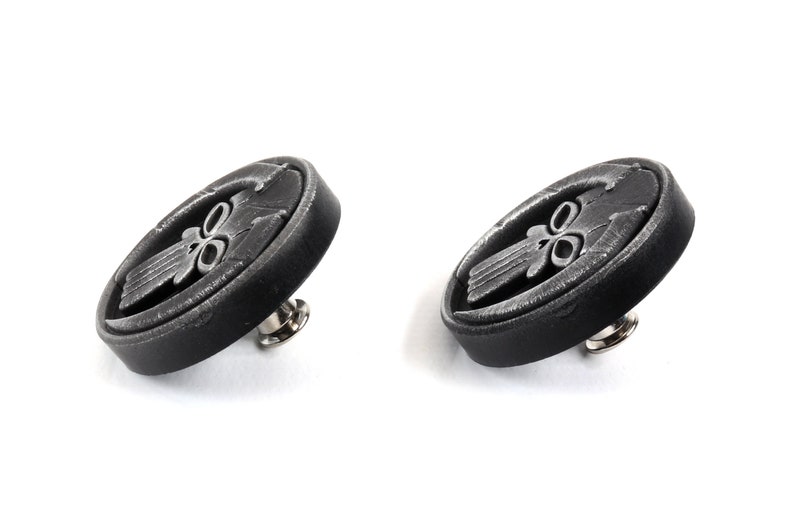 May include: Two black metal buttons with a silver skull design. Each button has a pin on the back.