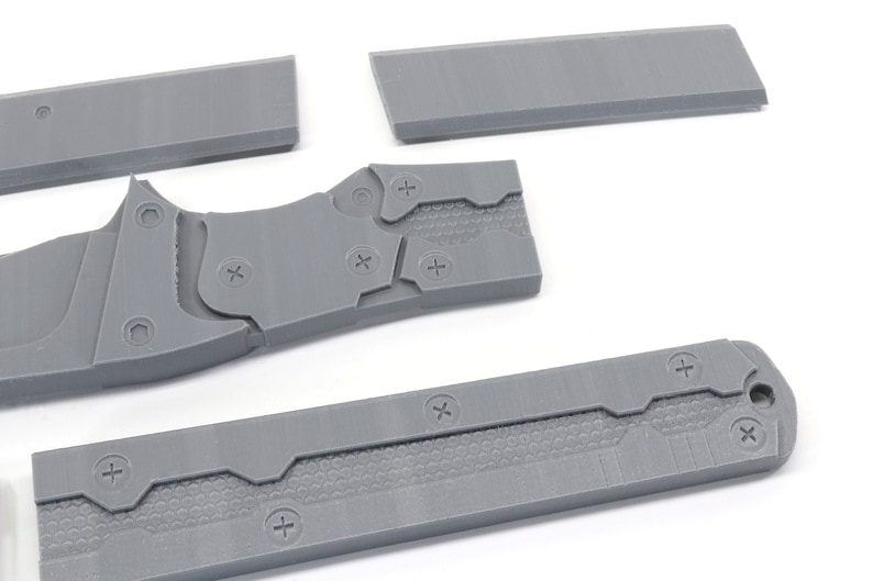 May include: Five gray plastic pieces of a disassembled knife. The pieces have a textured surface and are held together by screws.