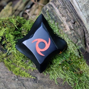 May include: Black ninja star with a red swirl design, resting on a mossy log.
