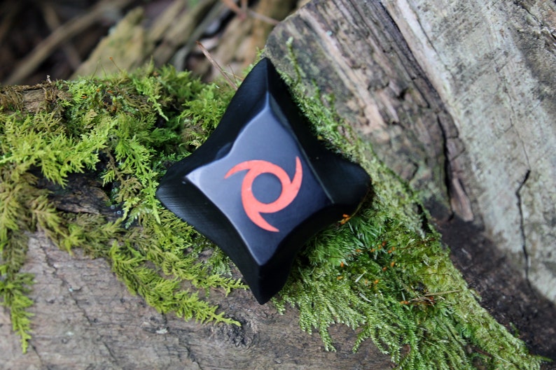 May include: Black ninja star with a red swirl design, resting on a mossy log.
