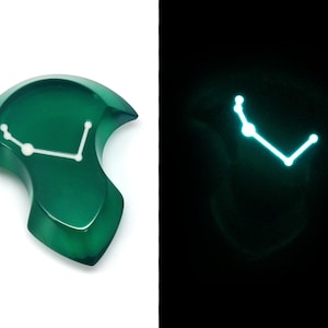 May include: A green, abstract-shaped pendant with a white constellation design. The pendant glows with a bright blue light in the dark. The pendant is on a white background.