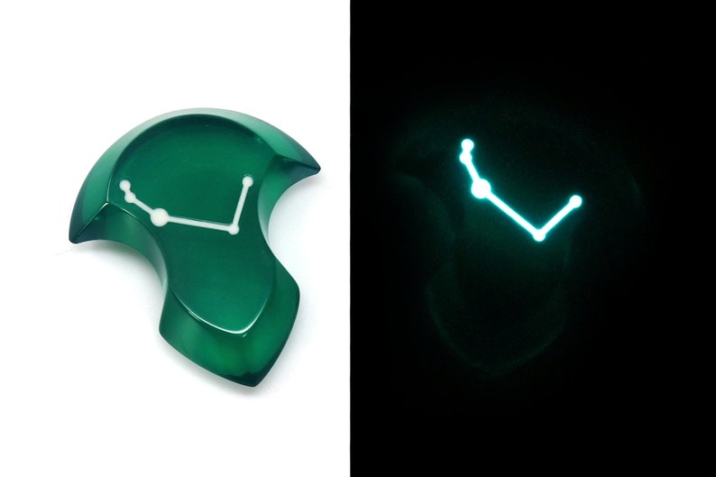 May include: A green, abstract-shaped pendant with a white constellation design. The pendant glows with a bright blue light in the dark. The pendant is on a white background.