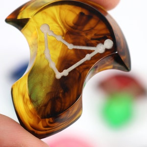 May include: A crescent-shaped, amber-coloured resin guitar pick with a white constellation design. The constellation is Pisces, the fish.